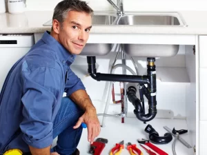 A plumber kneels beside an open cabinet under a kitchen sink in Toronto, surrounded by various plumbing tools, smiling at the camera while expertly handling a drain repair and plumbing project.