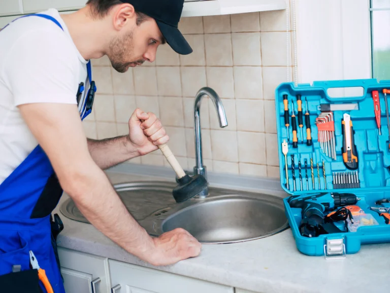 A plumber in a blue uniform performs drain repair on a kitchen sink in Toronto. An open tool kit with various tools sits on the counter. Tiled walls and a window are visible in the background, highlighting expert plumbing in Toronto.