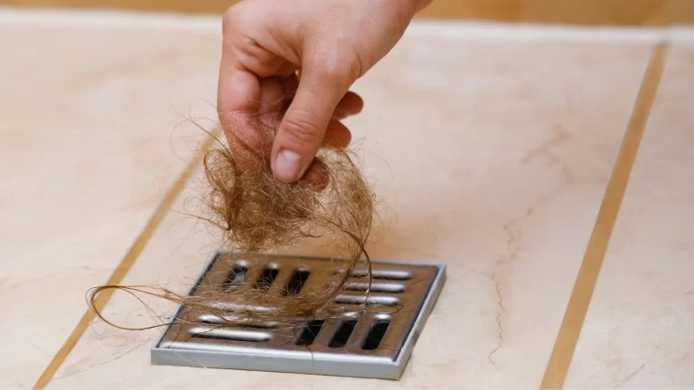 A hand picking up a clump of hair from a metal shower drain on a tiled bathroom floor.
