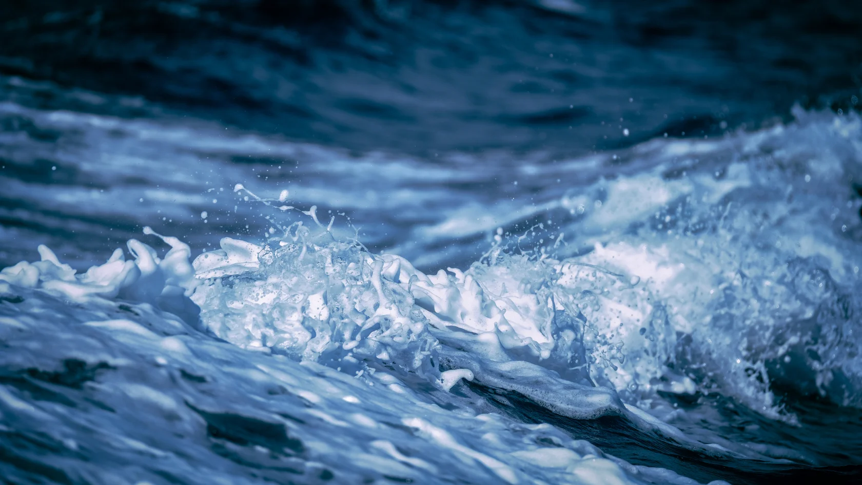 Close-up of ocean waves with white foam splashing on the surface, creating dynamic movement. The water appears deep blue, highlighting the turbulent and energetic nature of the sea.