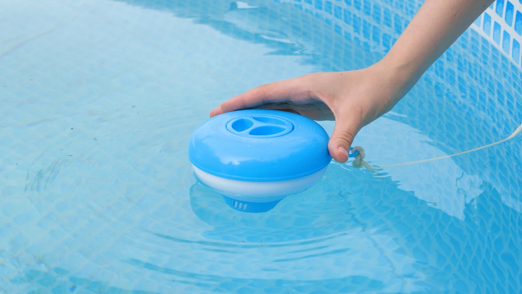 A hand holds a blue and white floating chlorine dispenser on the surface of a swimming pool with clear blue water and a tiled wall.