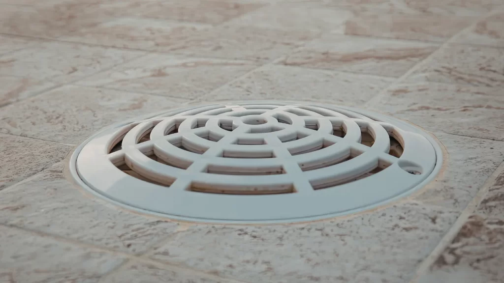 A close-up of a white, round drain cover with a grid pattern set into a tiled floor with light brown and beige tones.