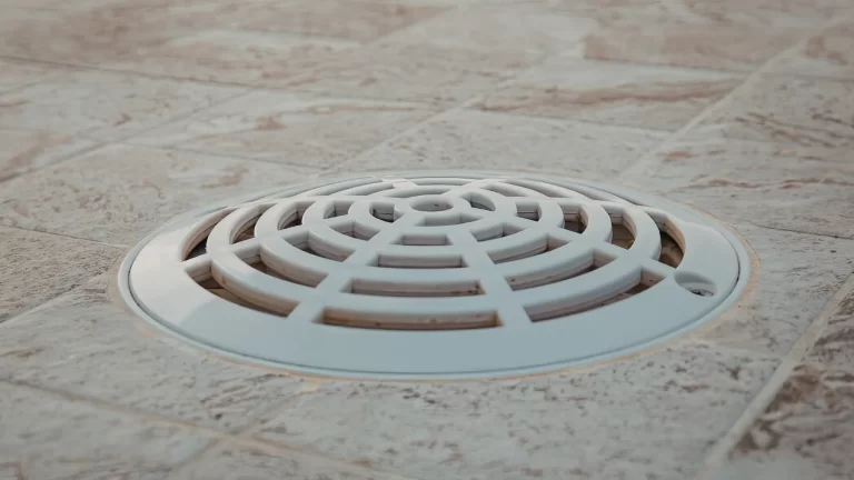 A close-up of a white, round drain cover with a grid pattern set into a tiled floor with light brown and beige tones.