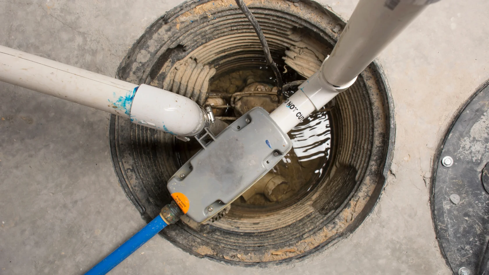 A close-up view of a sump pump system in a round pit, with several pipes connecting and water pooled at the bottom, set in a concrete floor.