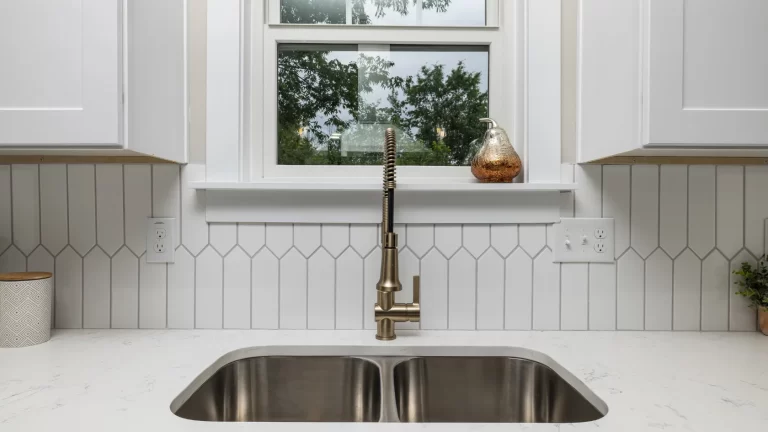 A modern kitchen sink with a double basin and a gold pull-down faucet is set in a white countertop. The backsplash features geometric white tiles, and a window above the sink shows green trees outside.