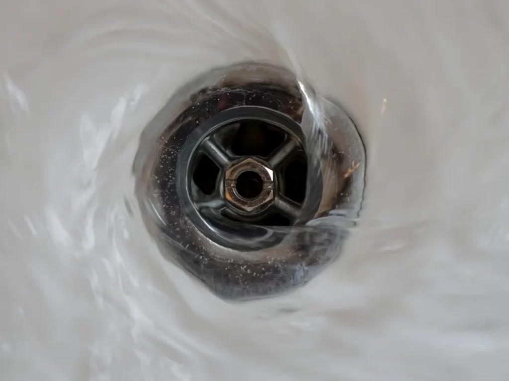 Close-up of water swirling around a metal drain in a white sink, creating a spiral motion as it flows down the drain.