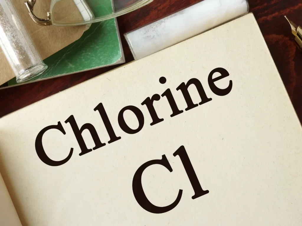 A close-up of a book or document page with the words “Chlorine Cl” printed in large, bold letters. Nearby are glass lab equipment and a green notebook, suggesting a scientific or educational context.