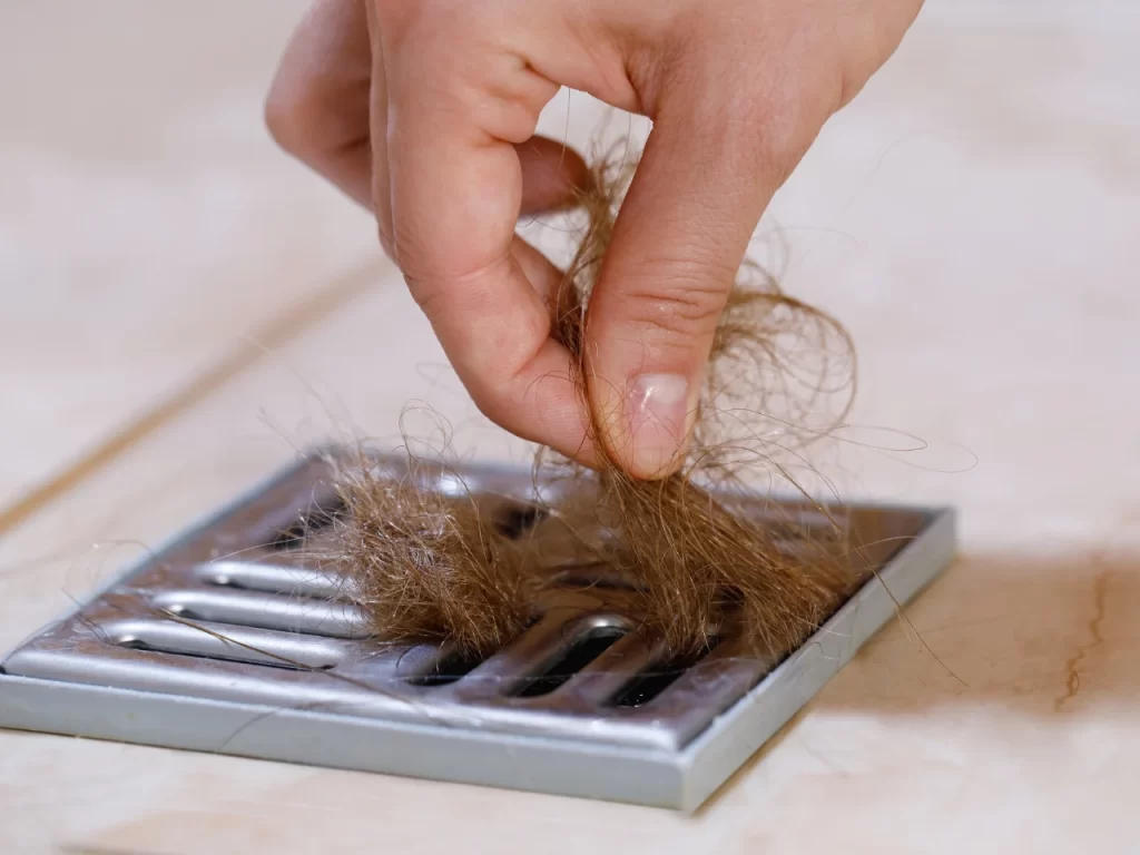 A hand is pulling a clump of brown hair out of a metal shower drain on a tiled floor.