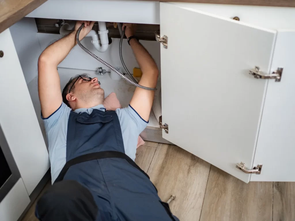 A plumber in a blue uniform lies on the floor under a kitchen sink, reaching up to work on the pipes inside an open cabinet. Tools and cleaning supplies are visible nearby.