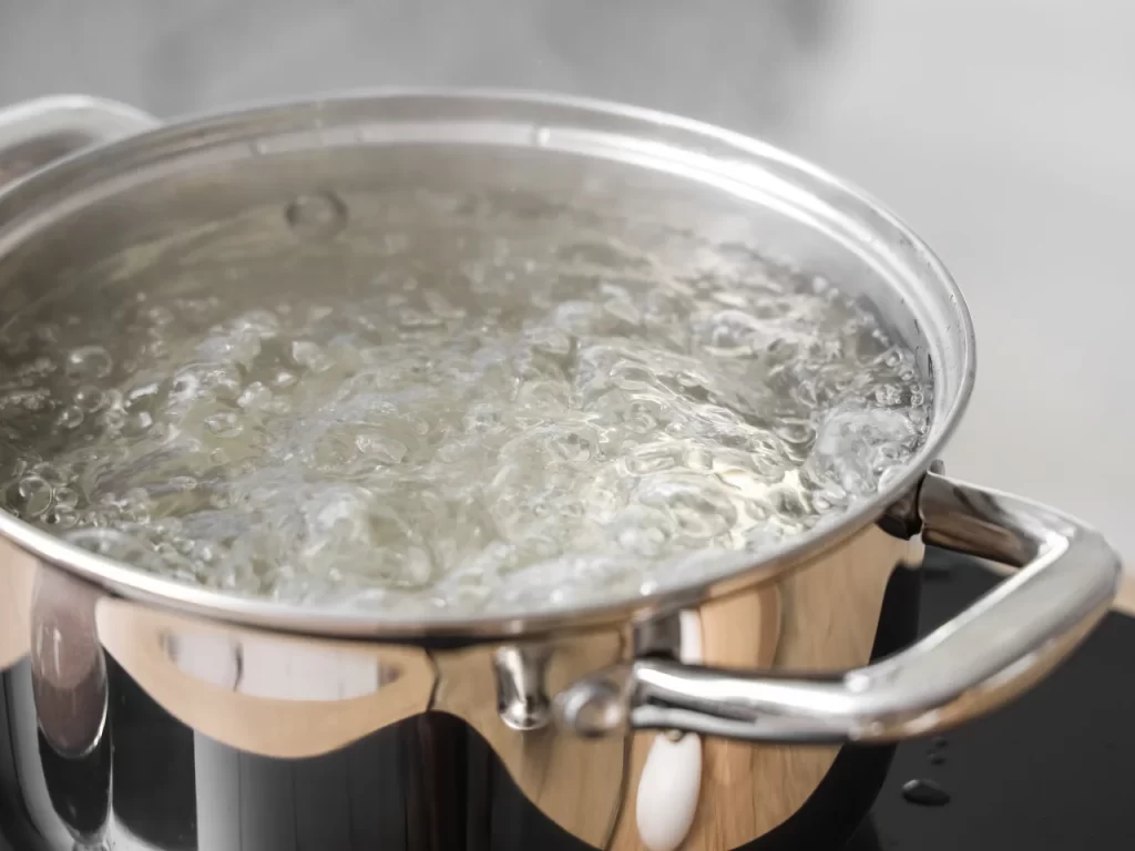 A stainless steel pot filled with water is boiling on a stovetop, with bubbles rising rapidly to the surface and steam visible above the pot.