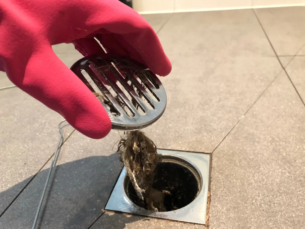 A person wearing a pink rubber glove lifts a metal drain cover from a floor drain, revealing a clump of hair and debris caught beneath it.