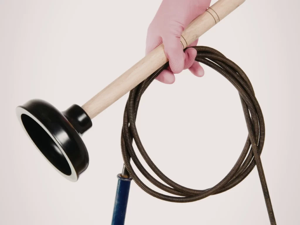 A hand wearing a pink glove holds a black plunger and a coiled plumbing snake against a plain white background.