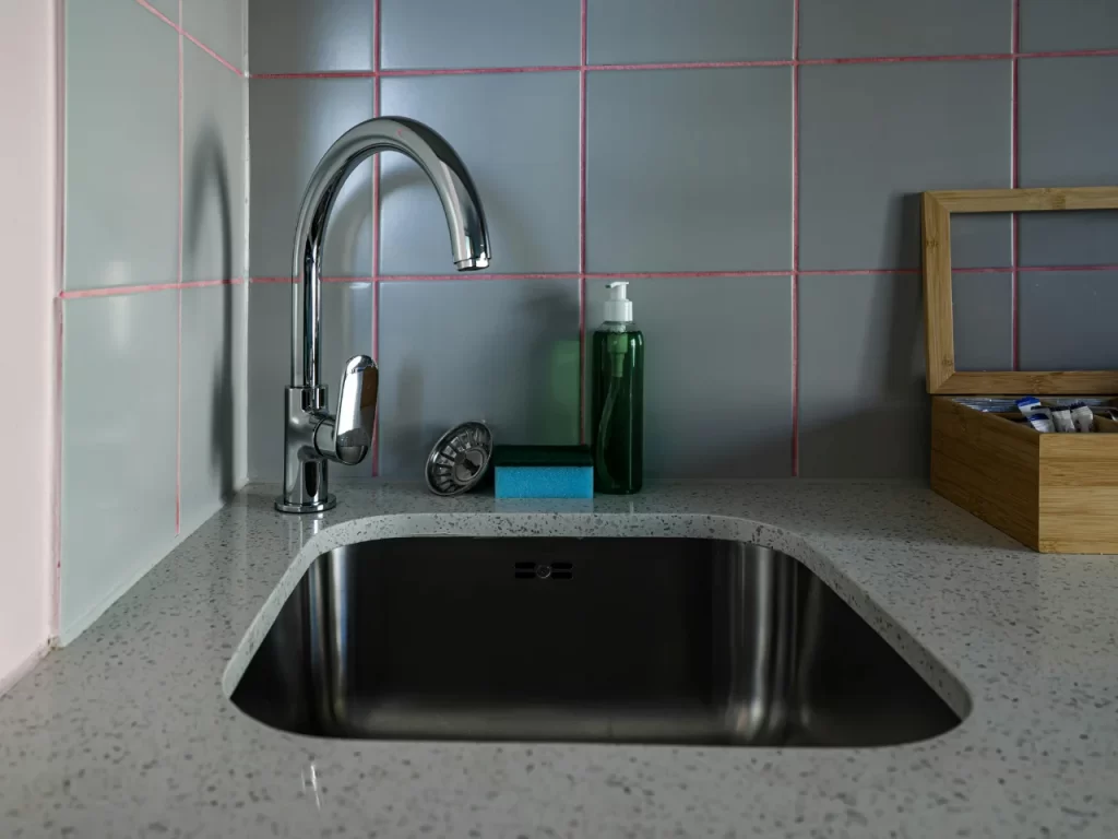 A modern kitchen sink with a curved faucet, soap dispenser, sponge, and a small timer on a speckled countertop. The backsplash features gray tiles with pink grout lines.