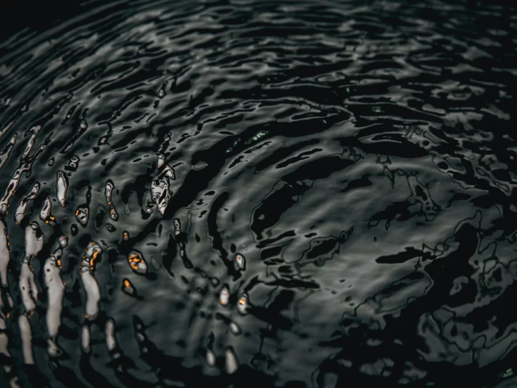 Dark, reflective ripples spread across the surface of water, creating abstract patterns and waves under low lighting. The image captures the texture and movement of the water with highlights and deep shadows.