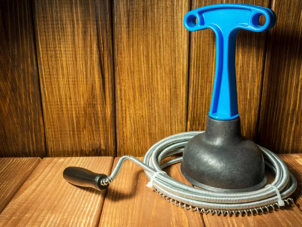 A plumbing snake with a blue handle and a coiled metal cable rests on a wooden surface with a wood-paneled background.