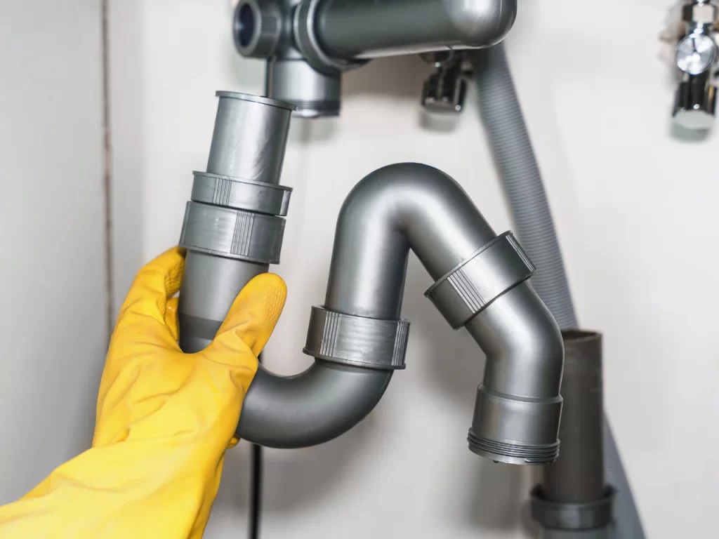 A person wearing a yellow rubber glove is holding a gray plastic P-trap pipe under a sink, with other plumbing components visible in the background.