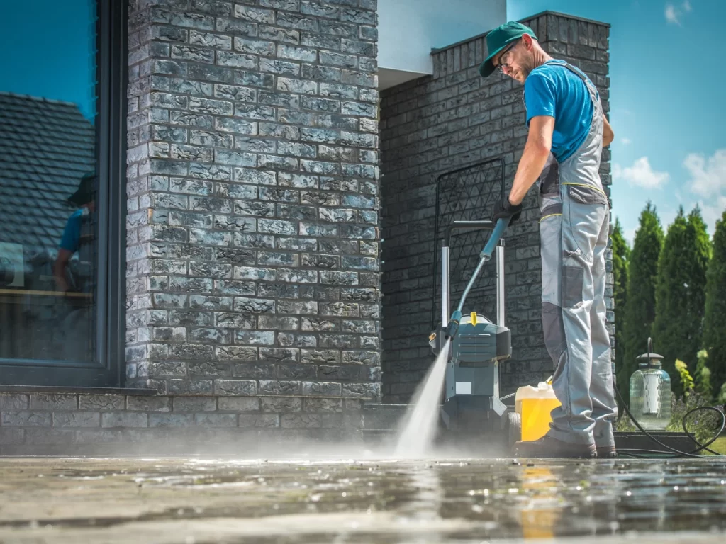 A man wearing work overalls and a cap uses a pressure washer to clean a paved patio area outside a modern brick house on a sunny day.