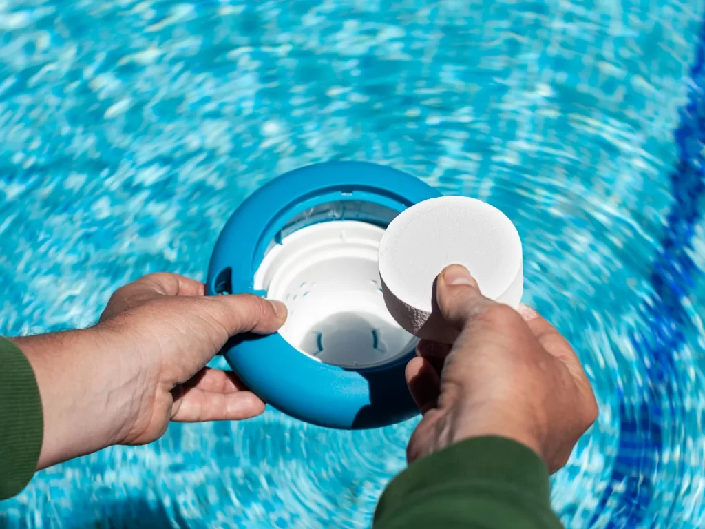 A person places a white chlorine tablet into a blue floating dispenser for a swimming pool, with clear water visible in the background.