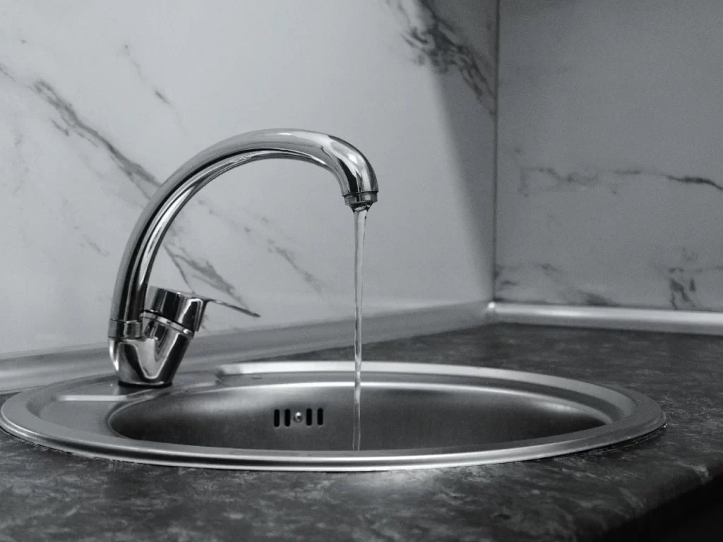 A modern chrome faucet releases a thin stream of water into a round, stainless steel sink set in a dark countertop, with a white and gray marble-patterned backsplash in the background.