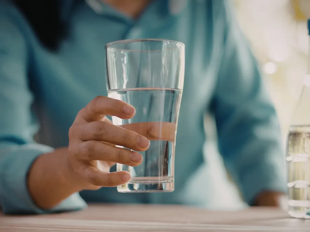 A person wearing a blue shirt holds a clear glass of water on a table, with natural light in the background.