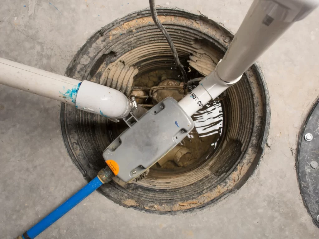 A close-up of a sump pump installed in a circular pit in a concrete floor, with several white PVC pipes and a blue hose connected, some water visible at the bottom of the pit.