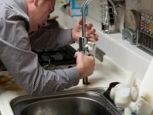 A person in a striped shirt is fixing or installing a kitchen faucet, using a tool to adjust the fixture above a stainless steel sink—showcasing expert drain repair and plumbing in Toronto.