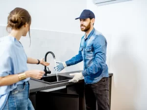 A plumber wearing a cap, gloves, and denim shirt is performing drain repair and plumbing in Toronto, installing or fixing a black kitchen faucet while a woman stands nearby, gesturing toward the sink.