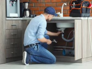 A plumber in a blue shirt, jeans, and cap is kneeling and working under a kitchen sink with tools, performing drain repair. A tool bag is on the counter, and the kitchen with wooden cabinets and a brick wall reflects quality plumbing in Toronto.