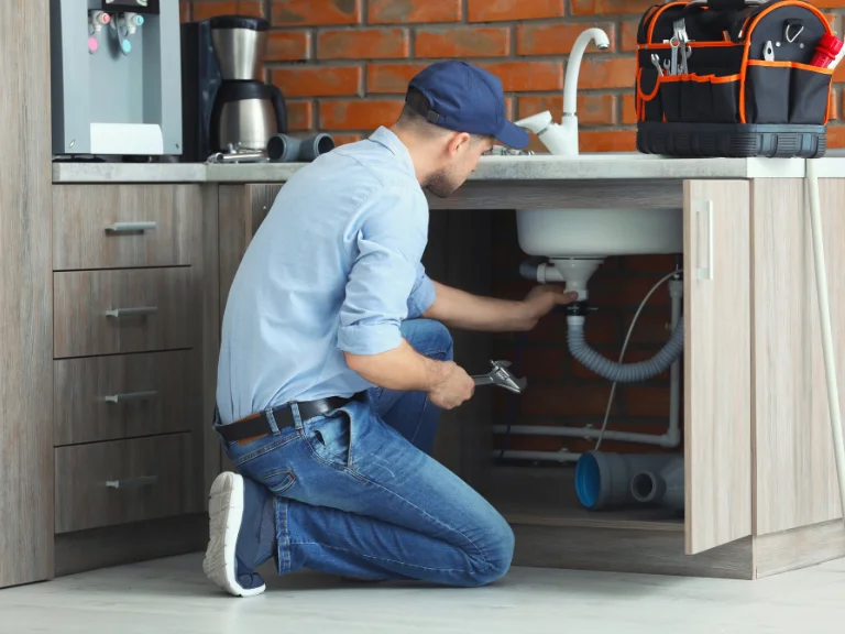 A plumber in a blue shirt, jeans, and cap is kneeling and working under a kitchen sink with tools, performing drain repair. A tool bag is on the counter, and the kitchen with wooden cabinets and a brick wall reflects quality plumbing in Toronto.