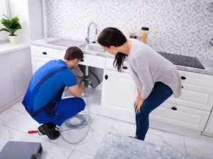 A plumber in blue overalls handles drain repair under a kitchen sink while a woman watches. Tools and a coiled cable are on the white tile floor in the modern kitchen, highlighting expert plumbing in Toronto.