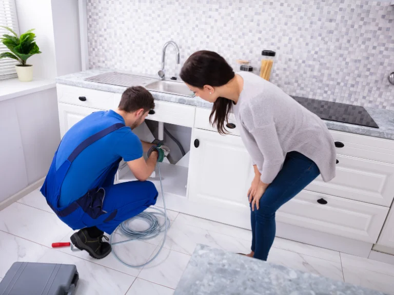 A plumber in blue overalls handles drain repair under a kitchen sink while a woman watches. Tools and a coiled cable are on the white tile floor in the modern kitchen, highlighting expert plumbing in Toronto.