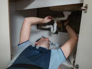 A plumber wearing glasses and a blue shirt is lying on his back under a sink, performing drain repair and plumbing in Toronto, using a tool to work on the white pipes inside a cabinet.