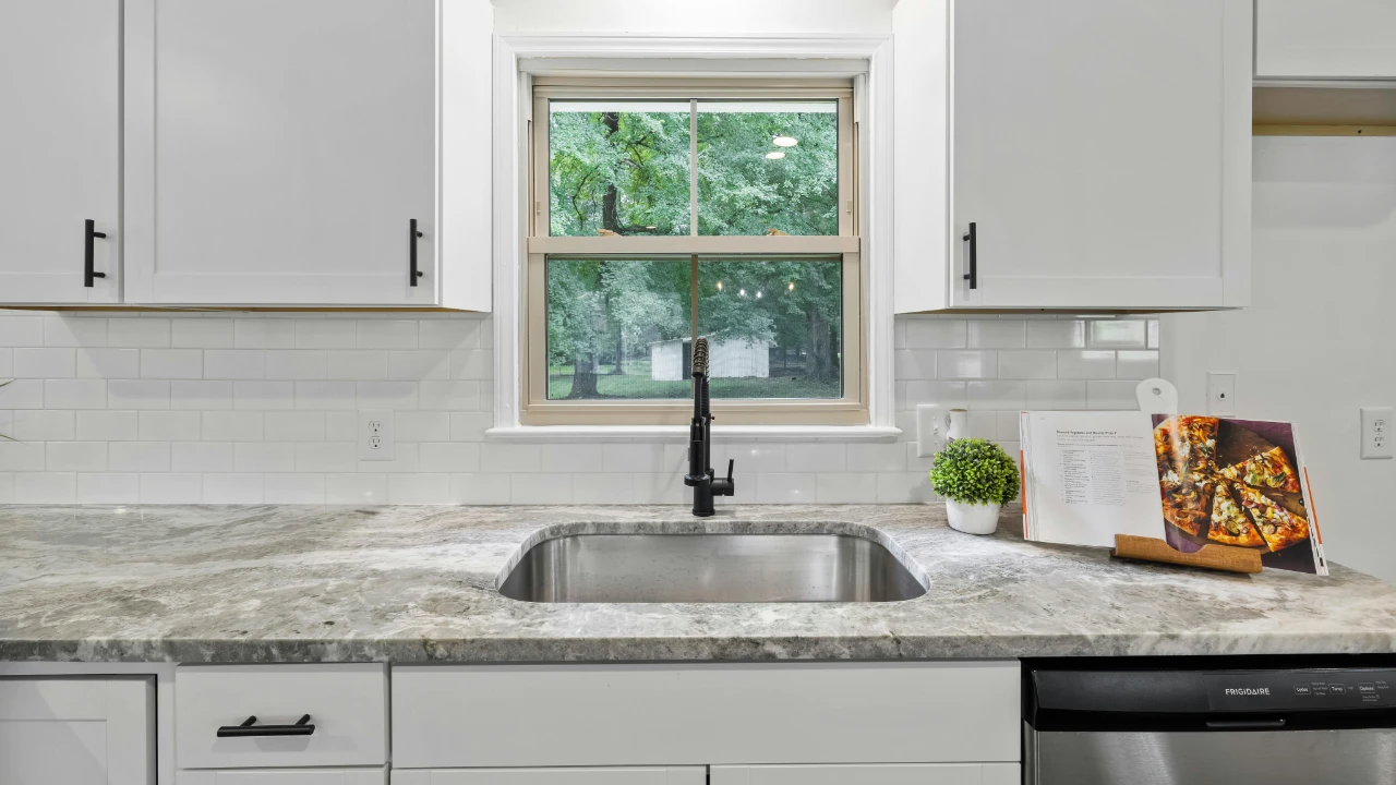 Modern kitchen with white cabinets, a stainless steel sink under a window, marble countertop, potted plant, paper towel holder, cookbook, and integrated dishwasher. Lush green trees are visible outside the window.