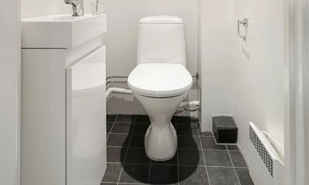 A small bathroom with a white toilet, white sink, and modern fixtures. The floor is covered with dark gray tiles and the walls are white. A toilet paper holder is mounted on the right wall.