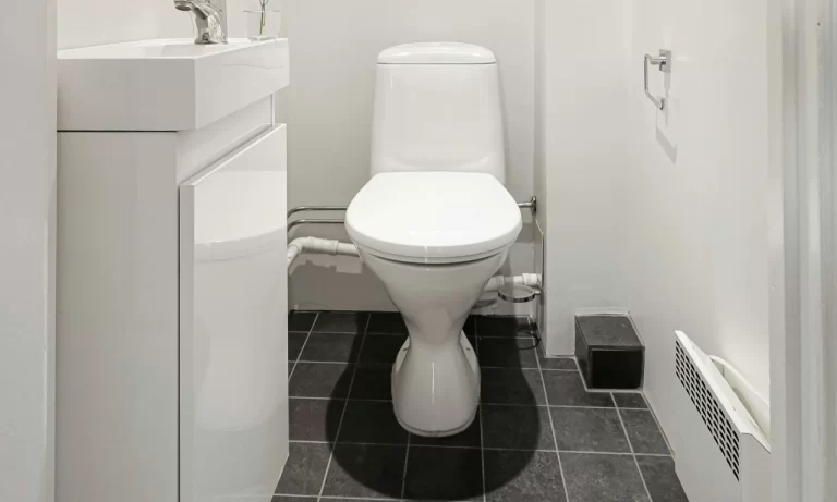 A small bathroom with a white toilet, white sink, and modern fixtures. The floor is covered with dark gray tiles and the walls are white. A toilet paper holder is mounted on the right wall.