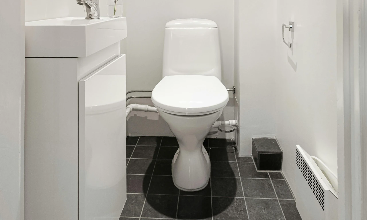 A small bathroom with a white toilet, white sink, and modern fixtures. The floor is covered with dark gray tiles and the walls are white. A toilet paper holder is mounted on the right wall.