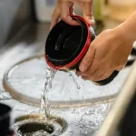 Hands are rinsing a film developing tank under running water in a sink, with water pouring out of the tank and splashing into the sink below.