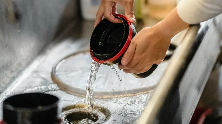 Hands are rinsing a film developing tank under running water in a sink, with water pouring out of the tank and splashing into the sink below.