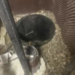 A person wearing a gray glove is working near a sump pit with exposed gravel, a submersible pump, black pipes, and utility equipment against a wall with dimpled brown waterproofing.