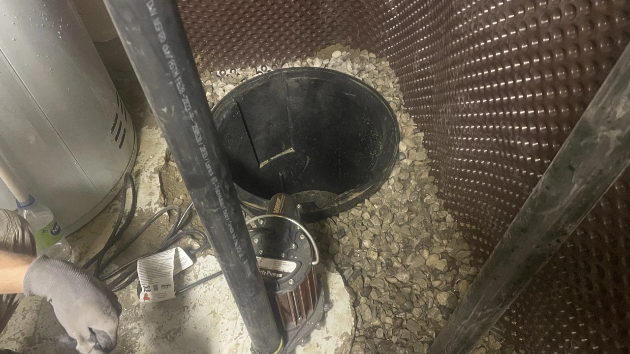 A person wearing a gray glove is working near a sump pit with exposed gravel, a submersible pump, black pipes, and utility equipment against a wall with dimpled brown waterproofing.