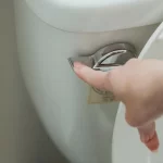 A hand is pressing down the silver flush lever on a white toilet, with the toilet lid partially raised.