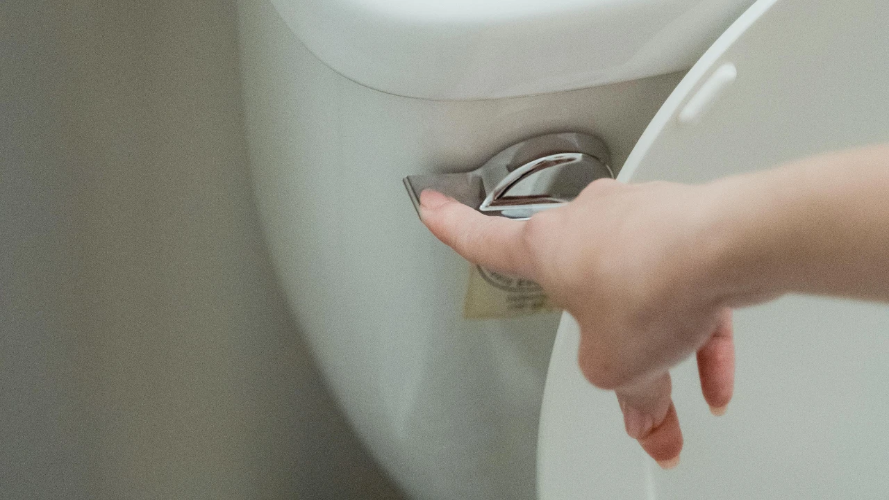A hand is pressing down the silver flush lever on a white toilet, with the toilet lid partially raised.