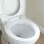 A clean white toilet with the lid and seat raised, positioned against a light-colored wall on a tiled bathroom floor.