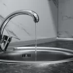 A modern metal kitchen faucet with a thin stream of water running into a round stainless steel sink, set against a marble-patterned backsplash and dark countertop.