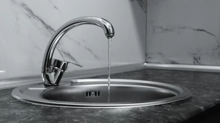 A modern metal kitchen faucet with a thin stream of water running into a round stainless steel sink, set against a marble-patterned backsplash and dark countertop.