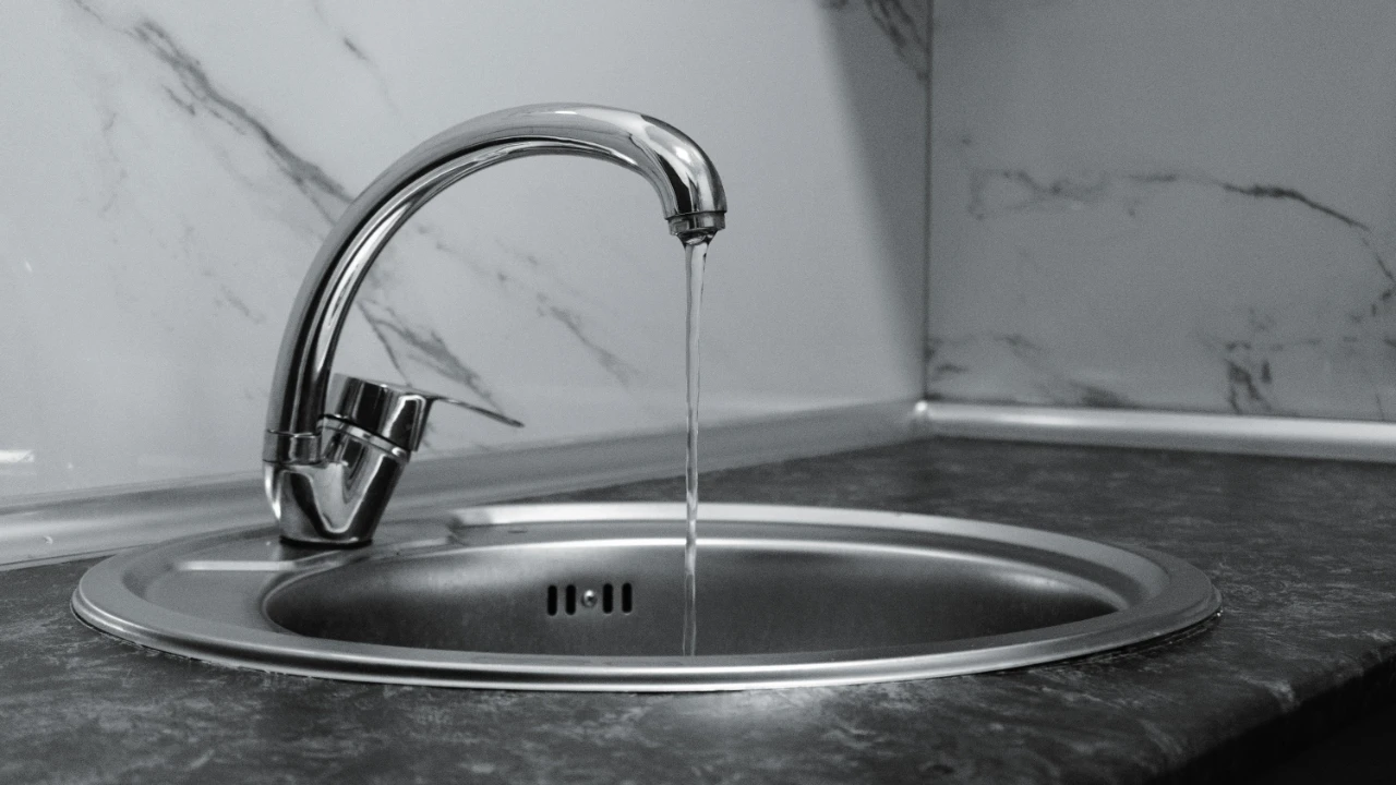 A modern metal kitchen faucet with a thin stream of water running into a round stainless steel sink, set against a marble-patterned backsplash and dark countertop.