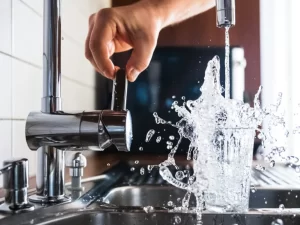 A hand adjusts a kitchen faucet while water overflows from a glass, splashing onto a metal sink—capturing dramatic droplets in mid-air, perfect for highlighting drain repair and plumbing in Toronto.