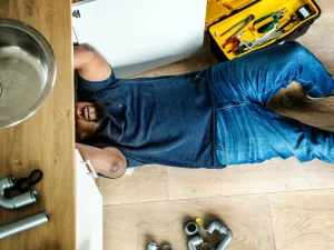 A man in a blue shirt and jeans lies on the wooden floor fixing plumbing under a sink, surrounded by a yellow toolbox and scattered pipes—typical of drain repair and plumbing in Toronto.