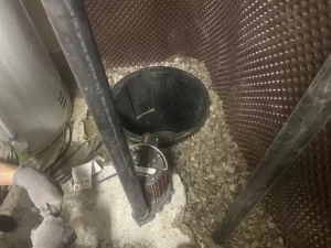 A person wearing gloves works next to an open sump pit surrounded by rocks and pipes in a basement or utility area, handling tools and equipment for drain repair and plumbing in Toronto.