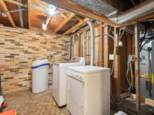 A basement laundry room with exposed wooden beams and pipes, a washing machine, dryer, water softener, and a brick-patterned wall. Some utility equipment and wiring are visible—ideal for those seeking expert plumbing in Toronto.